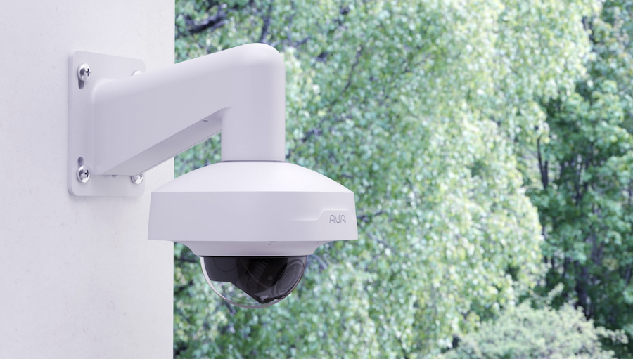 A white Ava CCTV security camera mounted on an exterior wall with trees in the background.